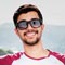 Smiling young man wearing sunglasses outdoors under a clear sky.